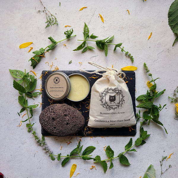 Open Tin of Hand & Foot Rescue balm, natural pumice stone, cotton bag of herbal foot soak, mint leaves and scattered flower petals