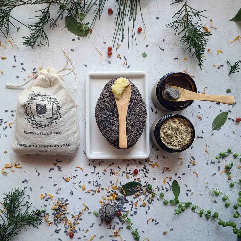 Texture, scooped Foot Balm and scooped Foot Mask. Open Foot Scrub, Lava Pumice and bag of Foot Soak. Scattered leaves and flowers.