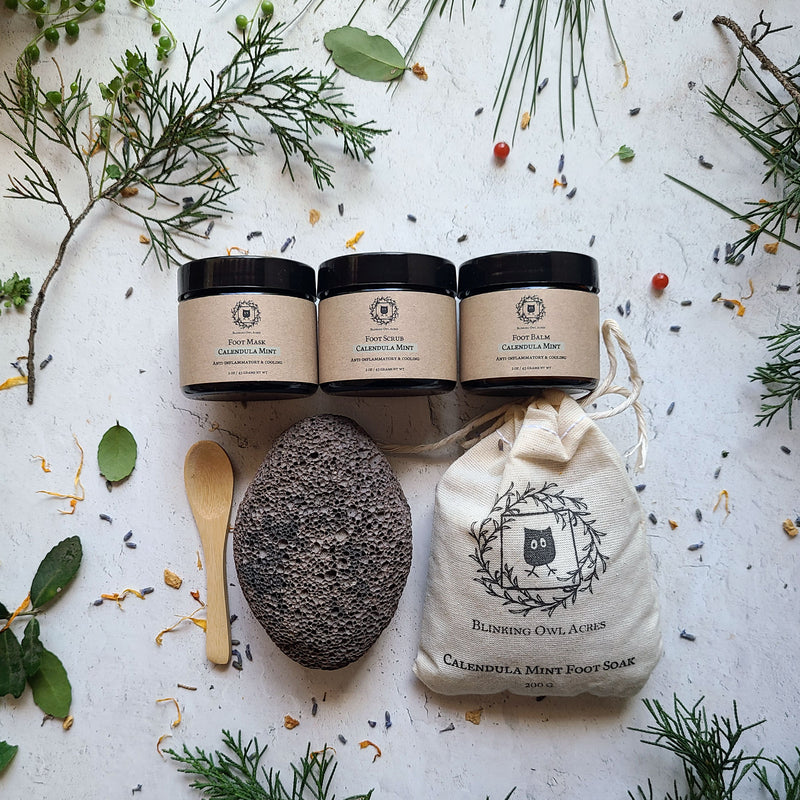 Three jars of calendula mint foot product, arranged with puice, foot soak bag and spoon, Botanical elements scattered decoratively.