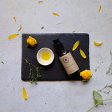 Treatment pump bottle of Herbal Oil Cleanse with oil in pinch pot and scattered botanical elements